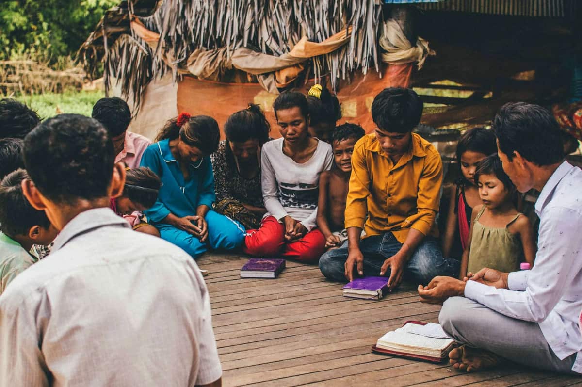 A local leader sits on a wooden floor with children and young adults as they read their Bibles together