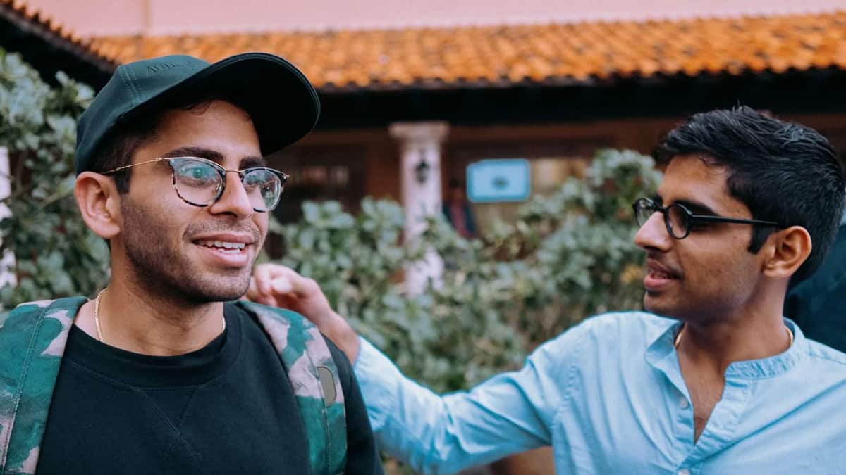 Two young men laugh together in a courtyard, one with a hand on the other's shoulder